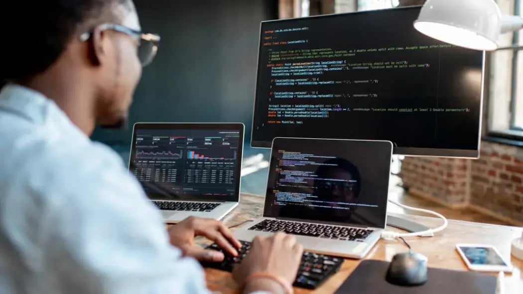 Man sat at desk with 3 computer screens all showing Python coding